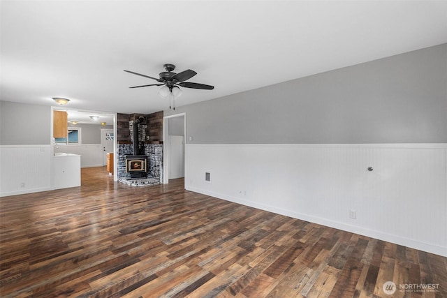 unfurnished living room with ceiling fan, a wood stove, wood finished floors, and wainscoting