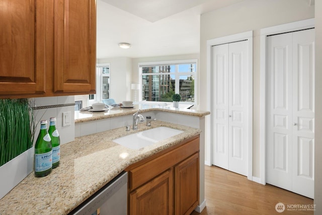 kitchen featuring brown cabinetry, light stone countertops, a sink, light wood-style floors, and stainless steel dishwasher