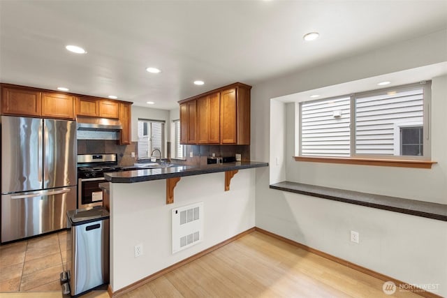 kitchen featuring brown cabinets, under cabinet range hood, backsplash, stainless steel appliances, and a peninsula