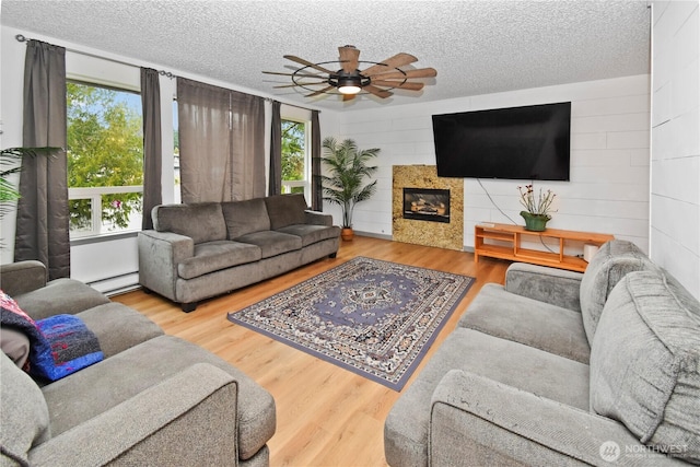 living room with ceiling fan, baseboard heating, wood finished floors, a glass covered fireplace, and a textured ceiling