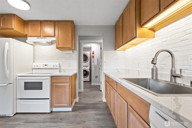 kitchen featuring under cabinet range hood, stacked washer and clothes dryer, wood finished floors, white appliances, and a sink