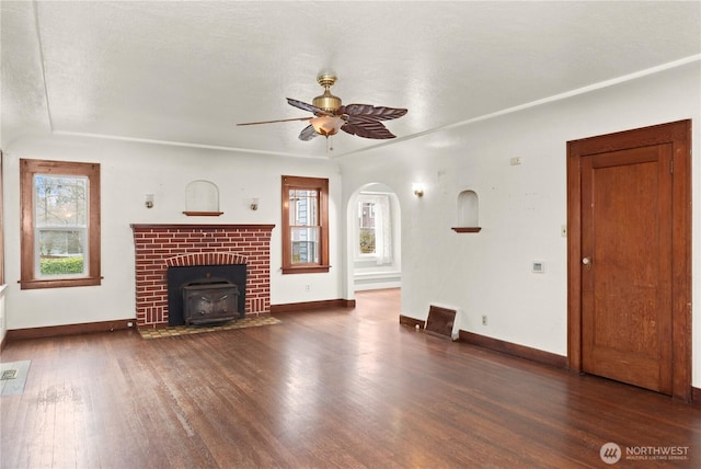 unfurnished living room featuring baseboards, a wood stove, wood finished floors, arched walkways, and a ceiling fan