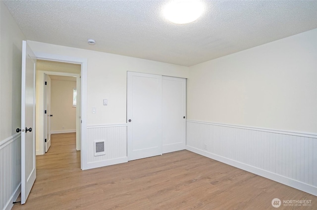 unfurnished bedroom featuring visible vents, a wainscoted wall, light wood-style flooring, a closet, and a textured ceiling