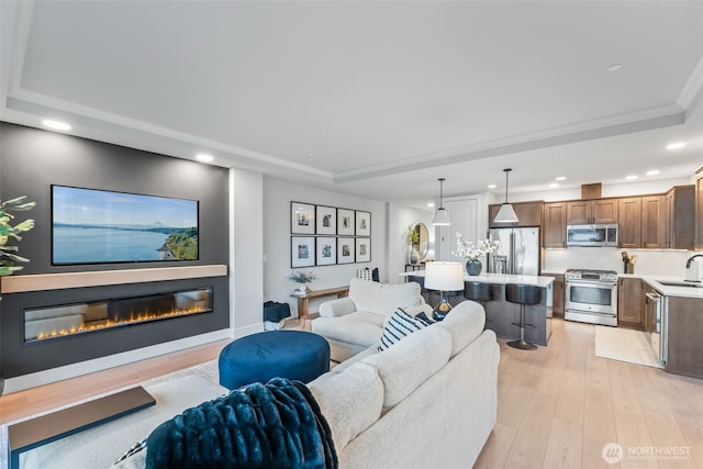 living area with a tray ceiling, light wood-style flooring, recessed lighting, a glass covered fireplace, and crown molding