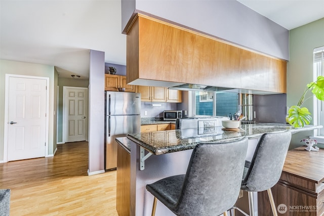 kitchen with a breakfast bar area, light wood-style flooring, dark stone countertops, brown cabinets, and stainless steel appliances