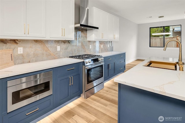 kitchen featuring stainless steel appliances, wall chimney range hood, a sink, blue cabinets, and light wood finished floors