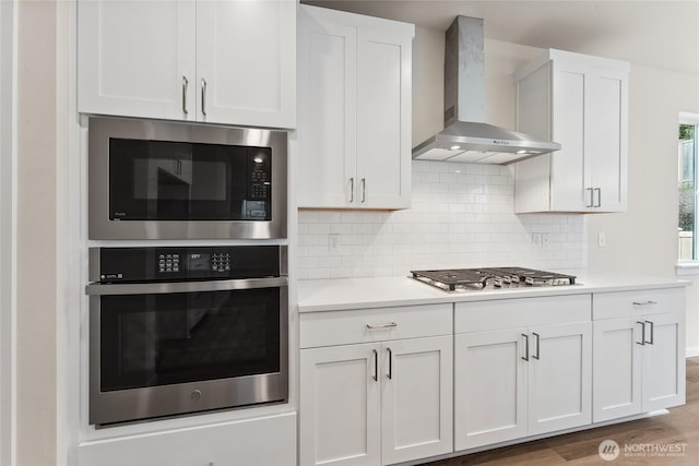 kitchen with wall chimney range hood, backsplash, stainless steel appliances, white cabinets, and light countertops