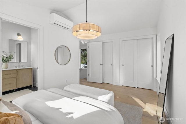 bedroom featuring connected bathroom, two closets, an AC wall unit, vaulted ceiling, and light wood-style flooring