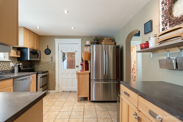 kitchen with stainless steel appliances, dark countertops, arched walkways, light tile patterned floors, and light brown cabinetry