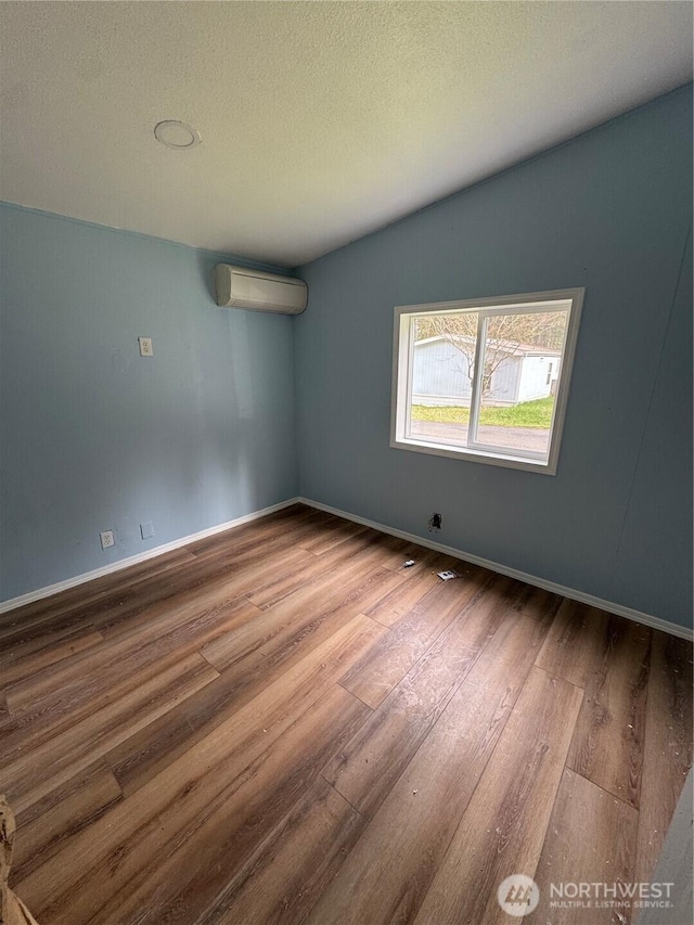 spare room featuring a wall mounted air conditioner, a textured ceiling, wood finished floors, baseboards, and vaulted ceiling