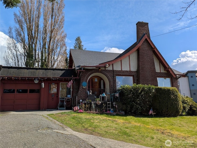 tudor home featuring roof with shingles, a front yard, an attached garage, brick siding, and a chimney