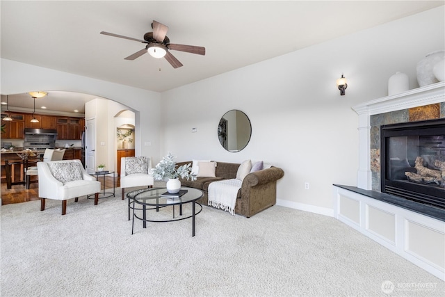 living room with baseboards, ceiling fan, a tiled fireplace, light colored carpet, and arched walkways