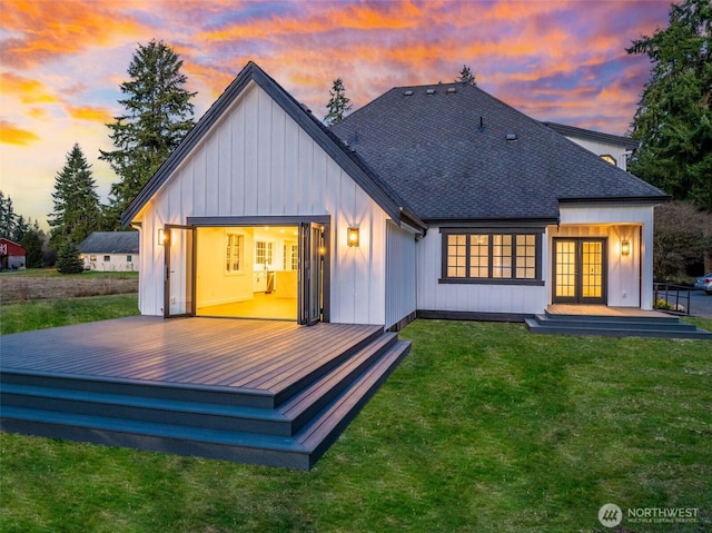 back of property at dusk featuring board and batten siding, a shingled roof, a wooden deck, french doors, and a yard