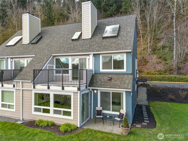 rear view of house featuring a yard, roof with shingles, a balcony, a chimney, and a patio area
