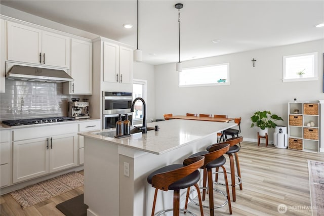 kitchen with light wood-style flooring, a kitchen breakfast bar, a sink, under cabinet range hood, and appliances with stainless steel finishes