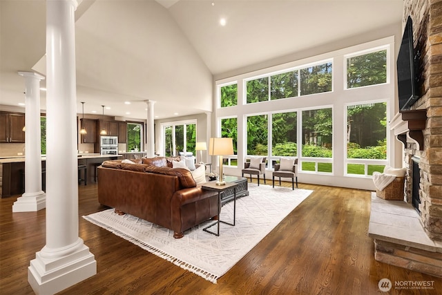 living area featuring high vaulted ceiling, a stone fireplace, dark wood-style flooring, and ornate columns