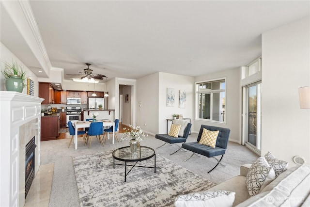 living area with baseboards, a fireplace with flush hearth, ceiling fan, crown molding, and light colored carpet