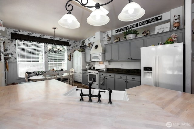 kitchen with custom range hood, wallpapered walls, an inviting chandelier, and white appliances