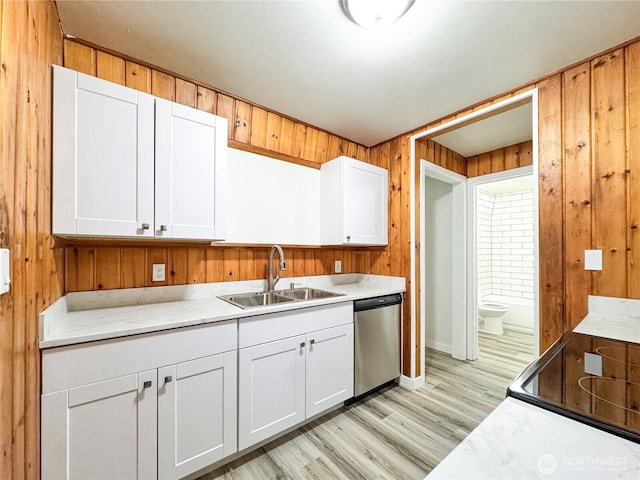 kitchen with a sink, dishwasher, light wood finished floors, white cabinets, and wood walls