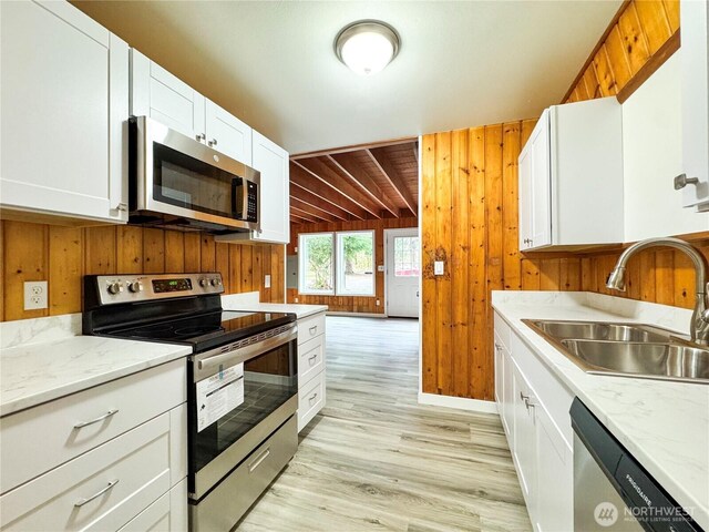 kitchen with a sink, light wood-type flooring, white cabinetry, wood walls, and appliances with stainless steel finishes