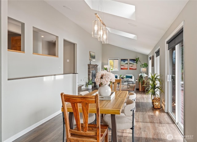 dining space featuring a stone fireplace, vaulted ceiling with skylight, baseboards, and wood finished floors