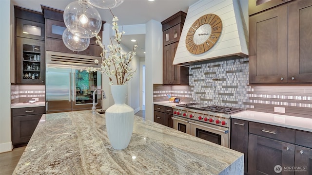 kitchen featuring dark brown cabinets, glass insert cabinets, premium range hood, high quality appliances, and decorative backsplash