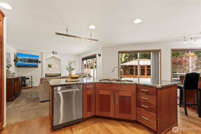 kitchen featuring stainless steel dishwasher, a sink, dark stone counters, open floor plan, and light wood-style floors