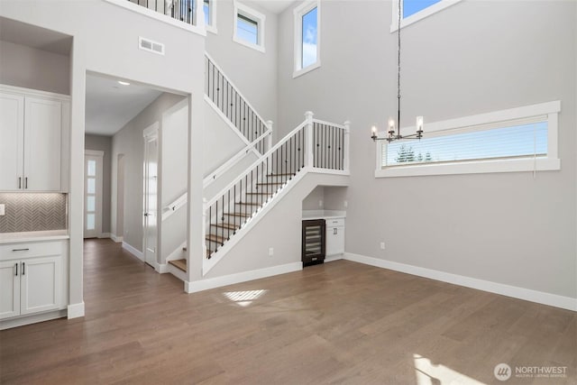 unfurnished living room with a towering ceiling, wood finished floors, stairway, a chandelier, and wine cooler