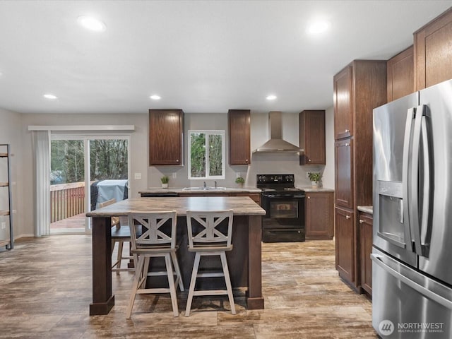kitchen with a kitchen bar, stainless steel fridge, black / electric stove, a sink, and wall chimney range hood