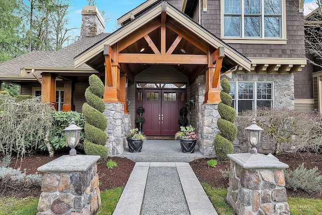 view of exterior entry featuring stone siding, a shingled roof, a chimney, and covered porch