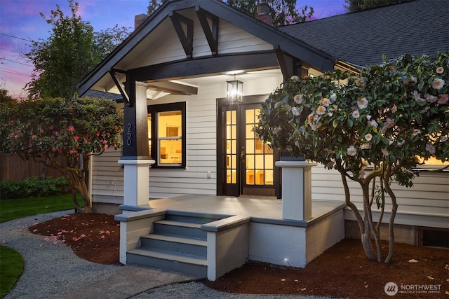 exterior entry at dusk with covered porch, a chimney, french doors, and roof with shingles
