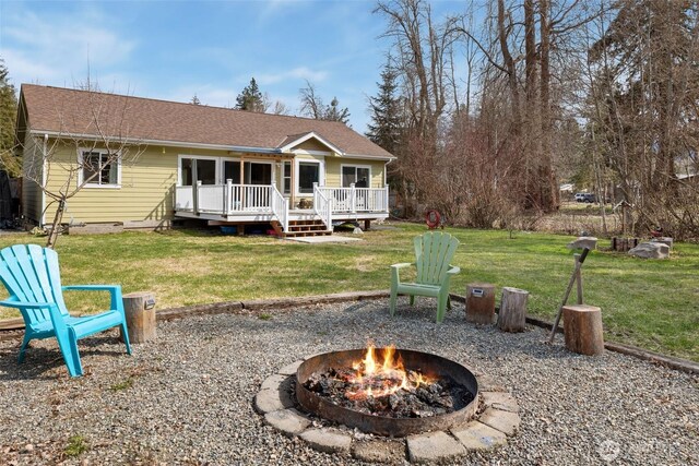rear view of property featuring a wooden deck, a fire pit, a lawn, and roof with shingles