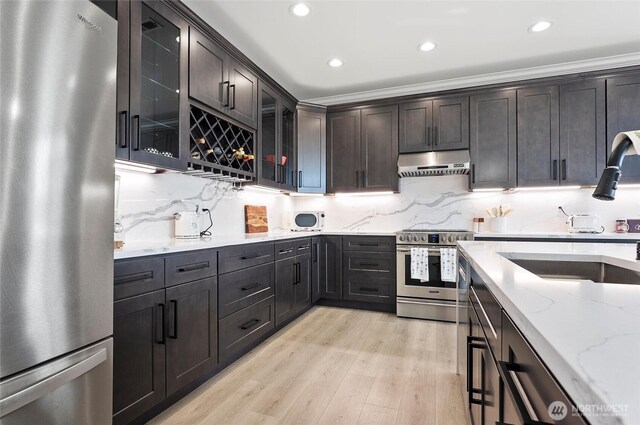 kitchen with stainless steel appliances, under cabinet range hood, a sink, light wood finished floors, and light stone countertops