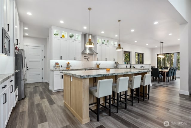 kitchen with an island with sink, built in microwave, wall chimney range hood, decorative backsplash, and dark wood-style flooring