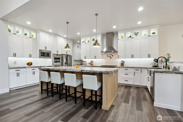 kitchen with dark wood-style flooring, stainless steel appliances, a sink, a kitchen island, and wall chimney exhaust hood