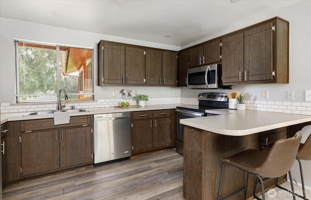 kitchen with a peninsula, wood finished floors, appliances with stainless steel finishes, dark brown cabinetry, and a sink