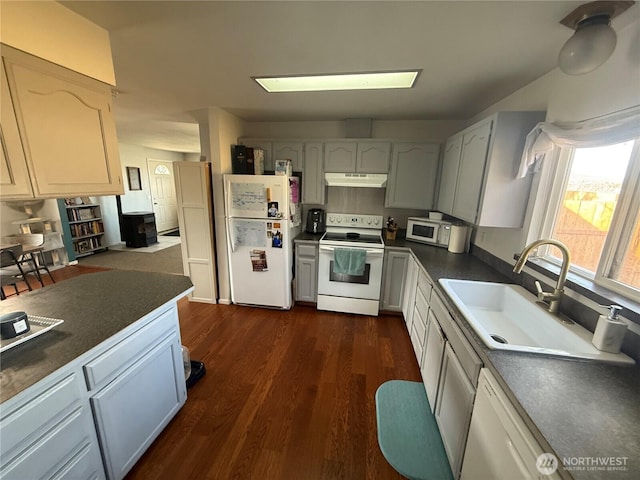 kitchen with a sink, under cabinet range hood, white appliances, dark countertops, and dark wood-type flooring