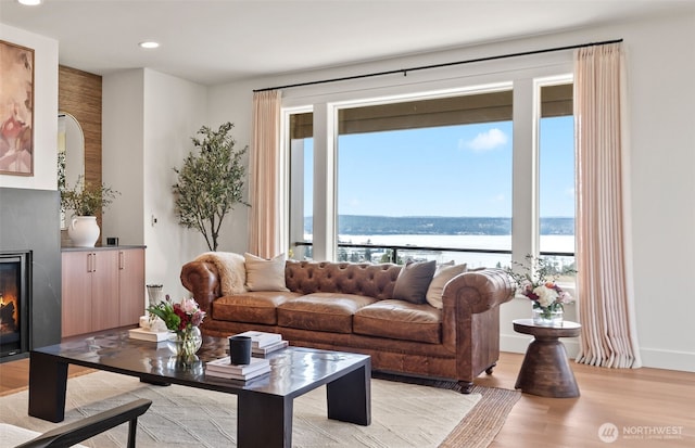living room featuring recessed lighting, light wood-type flooring, a water view, baseboards, and a glass covered fireplace
