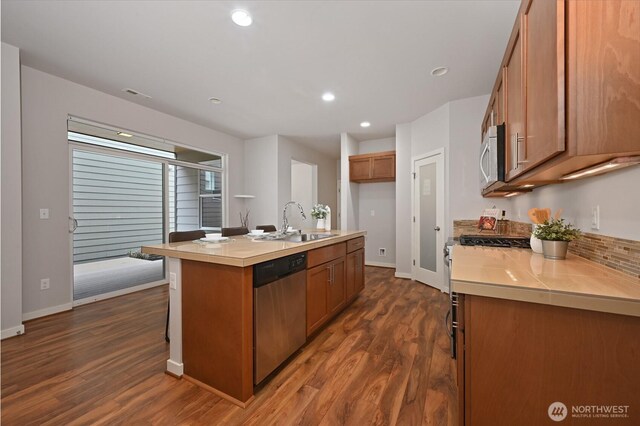 kitchen with light countertops, a sink, appliances with stainless steel finishes, brown cabinetry, and dark wood-type flooring