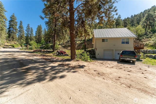 view of side of home with a garage, dirt driveway, a view of trees, metal roof, and a deck