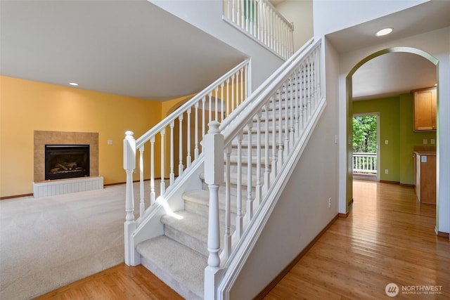 staircase with wood finished floors, baseboards, recessed lighting, a tiled fireplace, and arched walkways