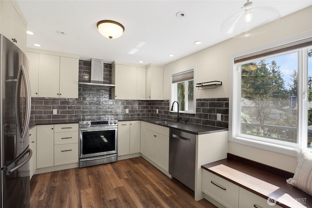 kitchen with stainless steel appliances, dark countertops, dark wood-type flooring, a sink, and wall chimney range hood