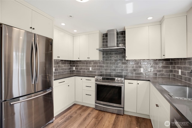 kitchen featuring a sink, light wood finished floors, appliances with stainless steel finishes, wall chimney range hood, and dark countertops