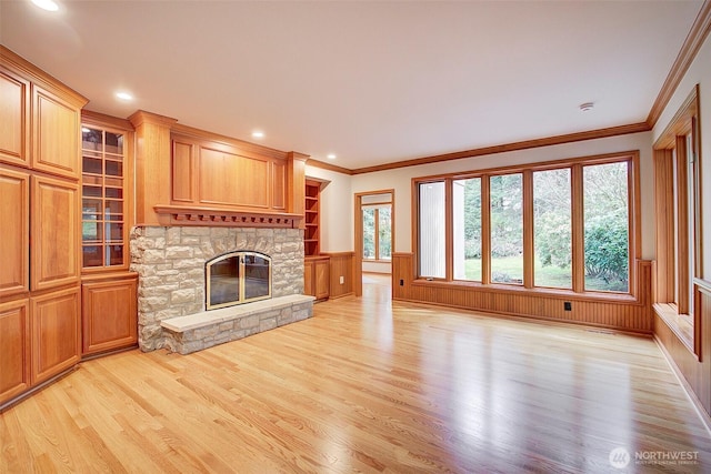 unfurnished living room featuring light wood-style floors, crown molding, a stone fireplace, recessed lighting, and wainscoting