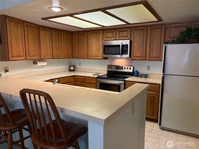 kitchen with stainless steel appliances, brown cabinetry, a kitchen bar, a peninsula, and light countertops