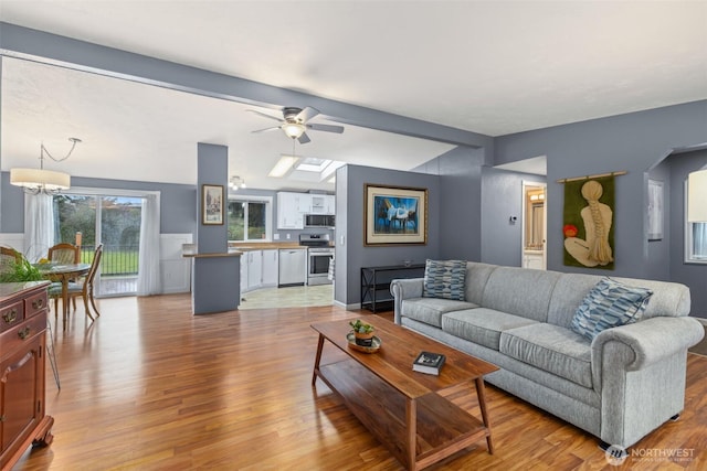 living room featuring light wood-style flooring, ceiling fan with notable chandelier, a wainscoted wall, and lofted ceiling with beams