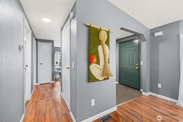 hallway with wood finished floors, a textured wall, baseboards, and visible vents