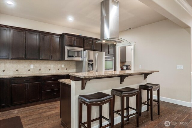 kitchen featuring dark wood-style flooring, island exhaust hood, a kitchen bar, stainless steel appliances, and tasteful backsplash