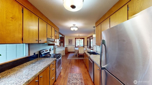 kitchen featuring a sink, wood finished floors, appliances with stainless steel finishes, brown cabinetry, and under cabinet range hood