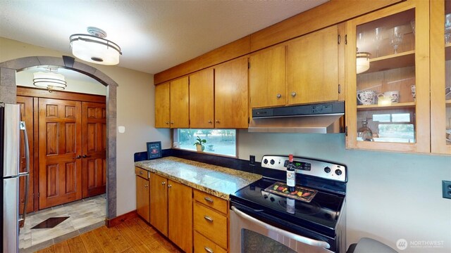 kitchen featuring light wood finished floors, appliances with stainless steel finishes, brown cabinets, under cabinet range hood, and arched walkways